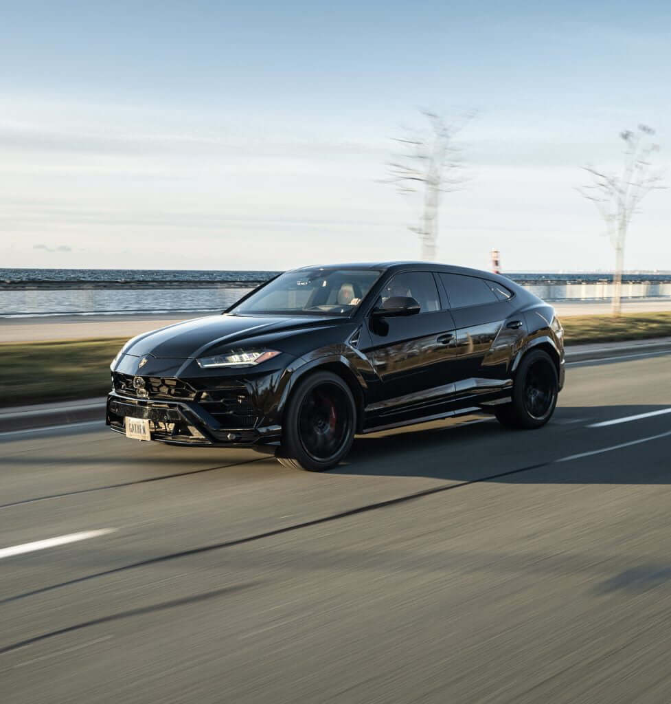 FX Productions Canada A black Lamborghini Urus SUV drives on a road near a waterfront on a clear day, showcasing the sleek style captured by FX Productions Canada, a leading Toronto Video Production company. Toronto Video Production Company