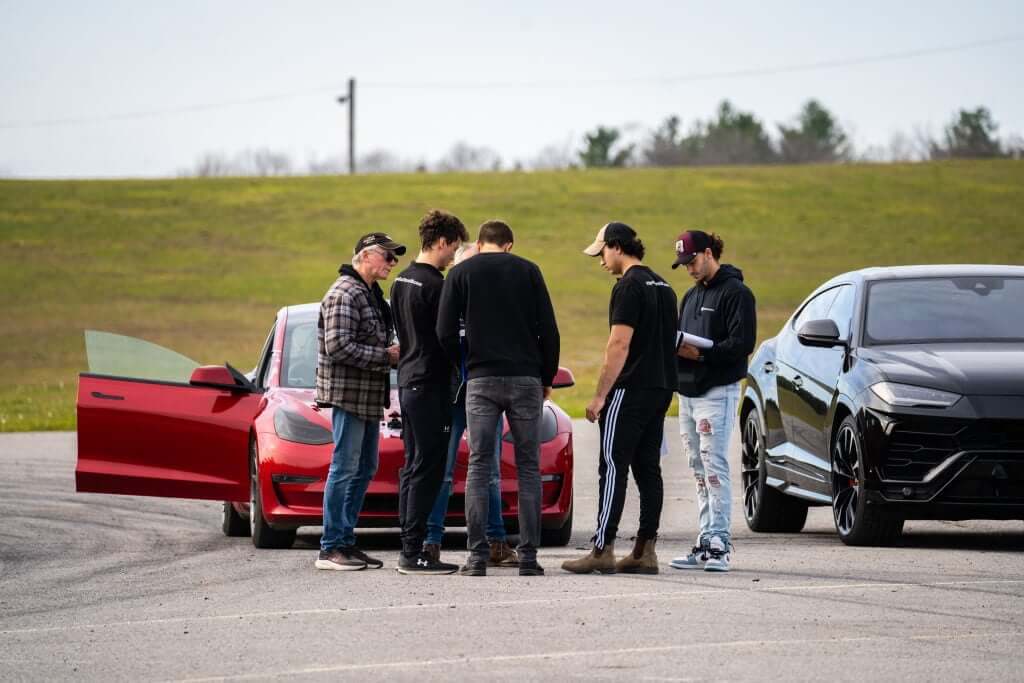 FX Productions Canada A group of six people stand between a red car with an open door and a black SUV in a parking lot with grassy hills in the background, during a Toronto Video Production shoot. Toronto Video Production Company
