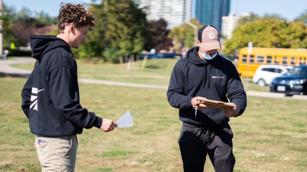 FX Productions Canada Two people stand outdoors on grass; one holds papers, the other writes on a clipboard. A yellow school bus and buildings are visible in the background, capturing a behind-the-scenes moment with FX Productions Canada, a Toronto Video Production company. Toronto Video Production Company