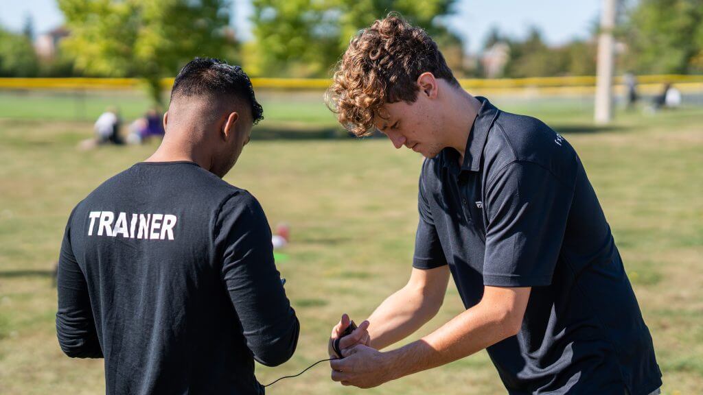 FX Productions Canada Two men standing on grass; one wears a shirt labeled "Trainer" while the other adjusts a device in his hands. Trees and a yellow fence are visible in the background—captured by FX Productions Canada, a leading Video Production Company. Toronto Video Production Company