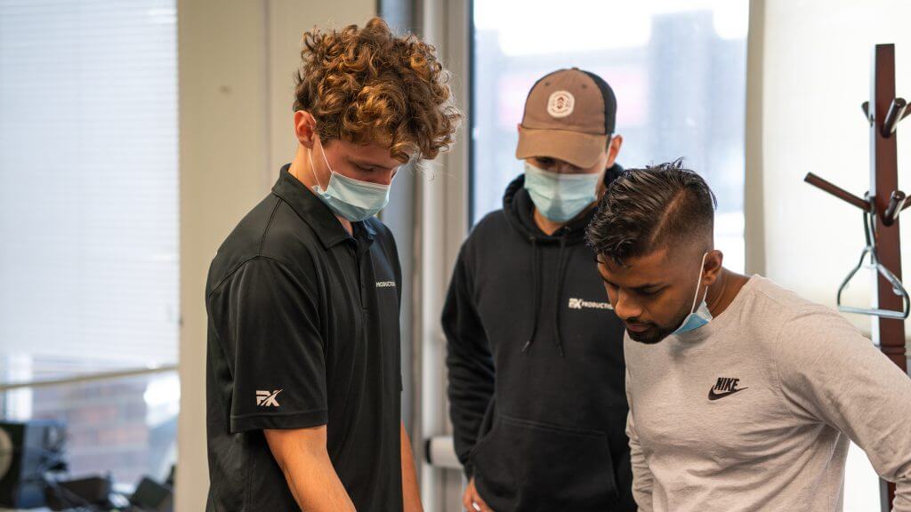 FX Productions Canada Three men wearing face masks and casual athletic clothing are standing indoors, closely observing something out of view at a Toronto Video Production shoot. Toronto Video Production Company