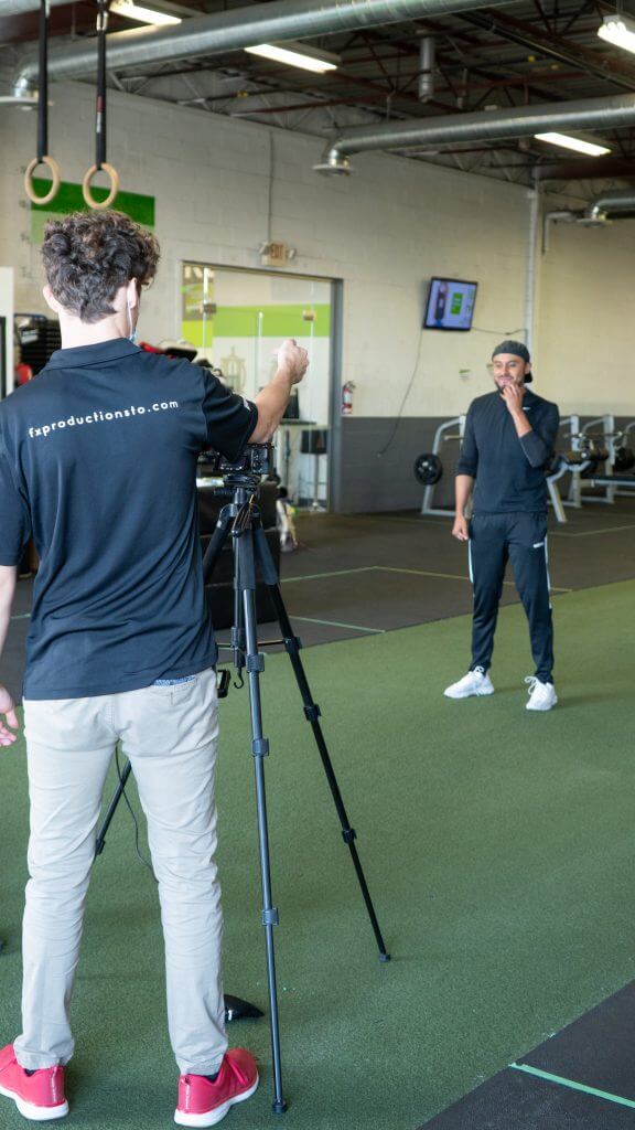 FX Productions Canada A man operates a camera on a tripod, filming another man standing on artificial turf in a gym with exercise equipment in the background—a behind-the-scenes look at Toronto Video Production in action. Toronto Video Production Company
