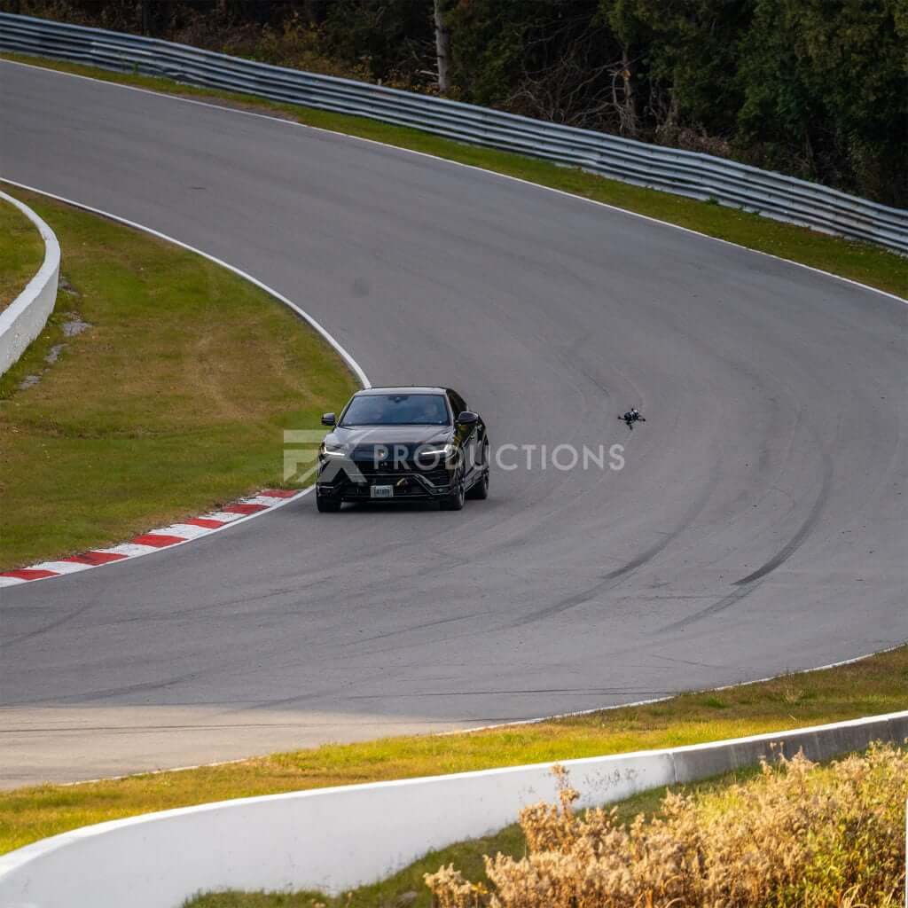 FX Productions Canada A black car speeds along a curving racetrack, closely followed by a drone. Trees and grass line the sides of the track, showcasing expert filming by FX Productions Canada. Toronto Video Production Company
