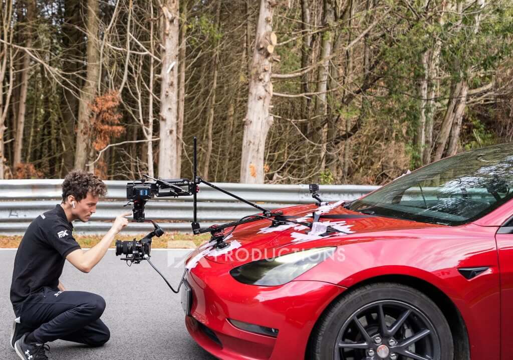 FX Productions Canada A person adjusts a camera rig mounted on the hood of a red car parked roadside near a forested area, capturing cinematic shots for FX Productions Canada. Toronto Video Production Company
