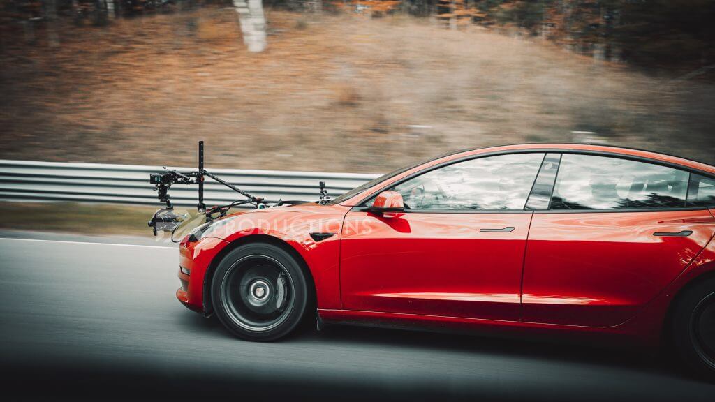 FX Productions Canada A red car with a camera rig mounted on the front drives on a road, captured by FX Productions Canada, with trees and a guardrail visible in the blurred background. Toronto Video Production Company