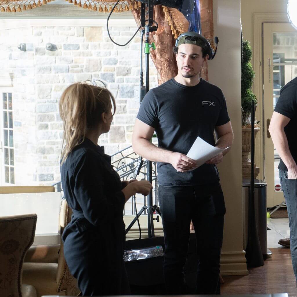 FX Productions Canada Two people stand indoors having a conversation; one holds papers and wears a black FX Productions Canada t-shirt and cap, while film equipment from a Toronto Video Production company is visible in the background. Toronto Video Production Company