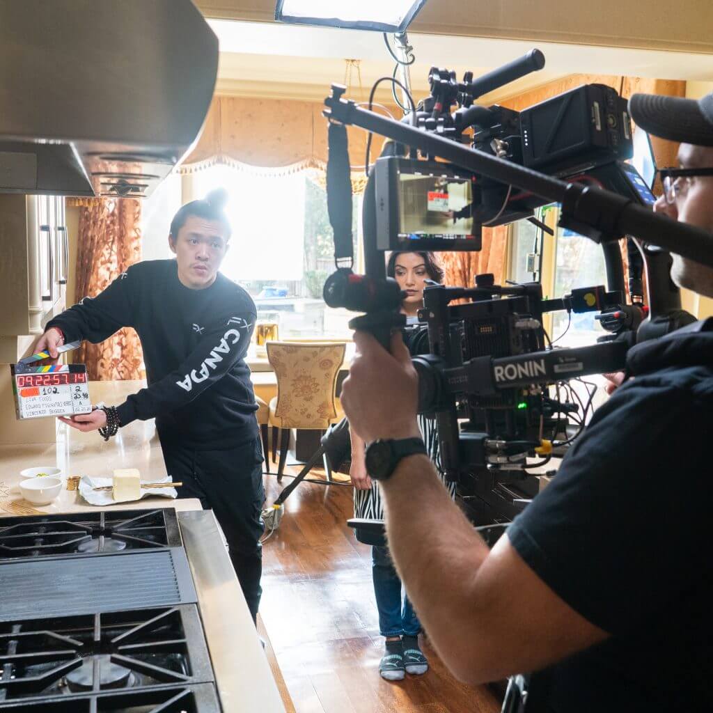 FX Productions Canada A Toronto Video Production crew films a scene in a kitchen, with a camera operator capturing a man holding a clapperboard and a woman standing beside him. Toronto Video Production Company