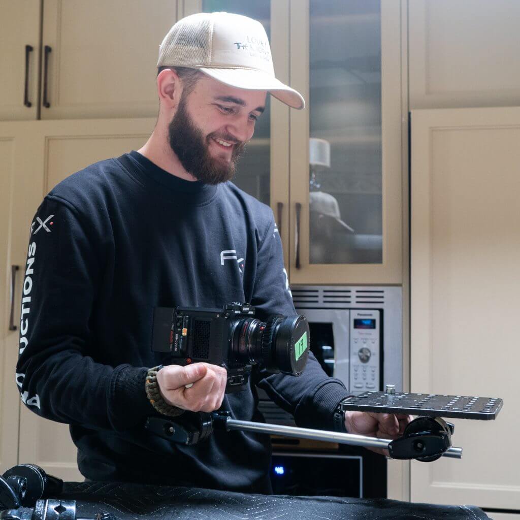 FX Productions Canada A man wearing a cap and dark sweatshirt holds a digital camera rig with a mounting plate in a kitchen setting, capturing behind-the-scenes moments for FX Productions Canada. Toronto Video Production Company