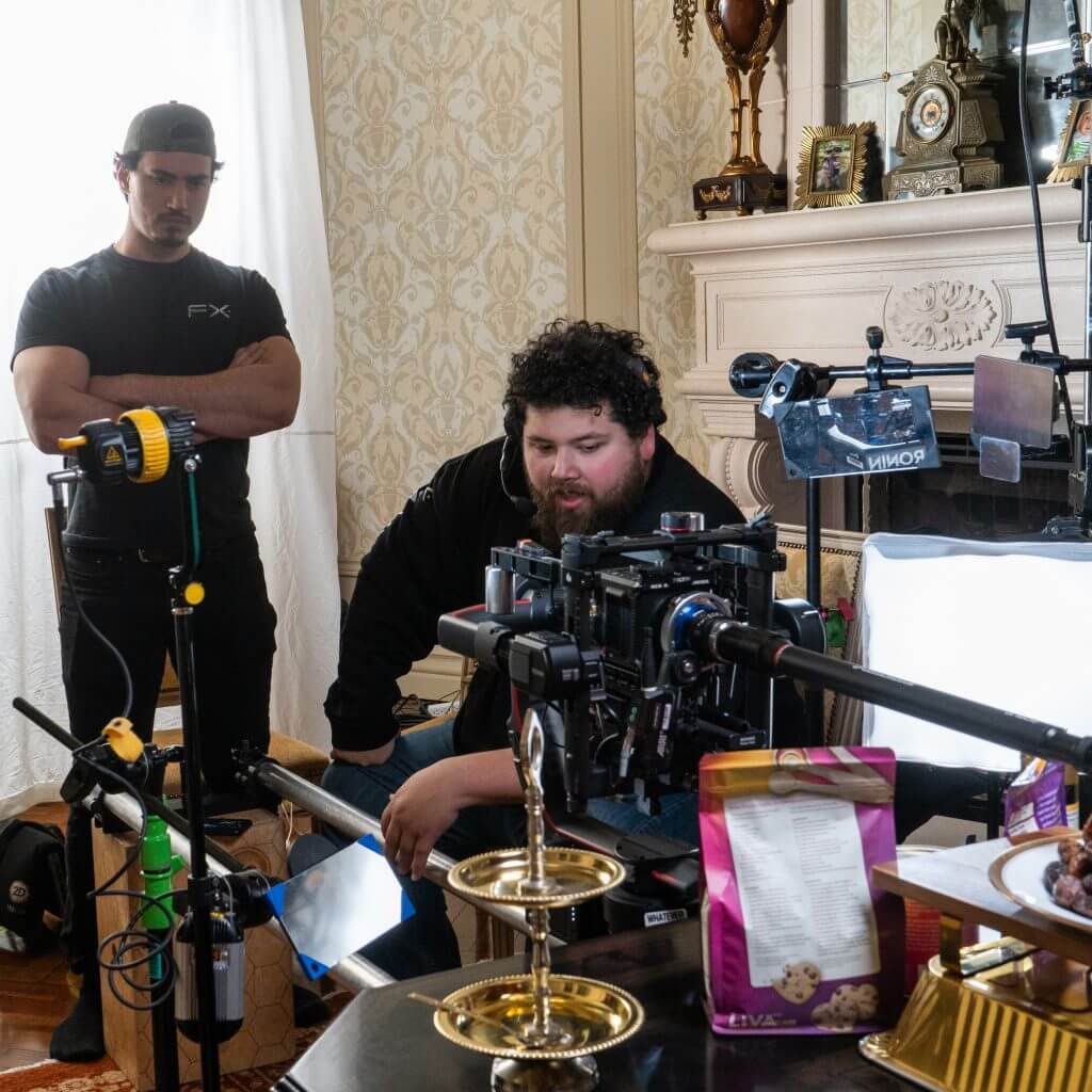 FX Productions Canada Two men stand behind a professional camera setup in a room with ornate wallpaper and a decorative fireplace; filming equipment and snack boxes are on the table, showcasing Toronto Video Production expertise. Toronto Video Production Company