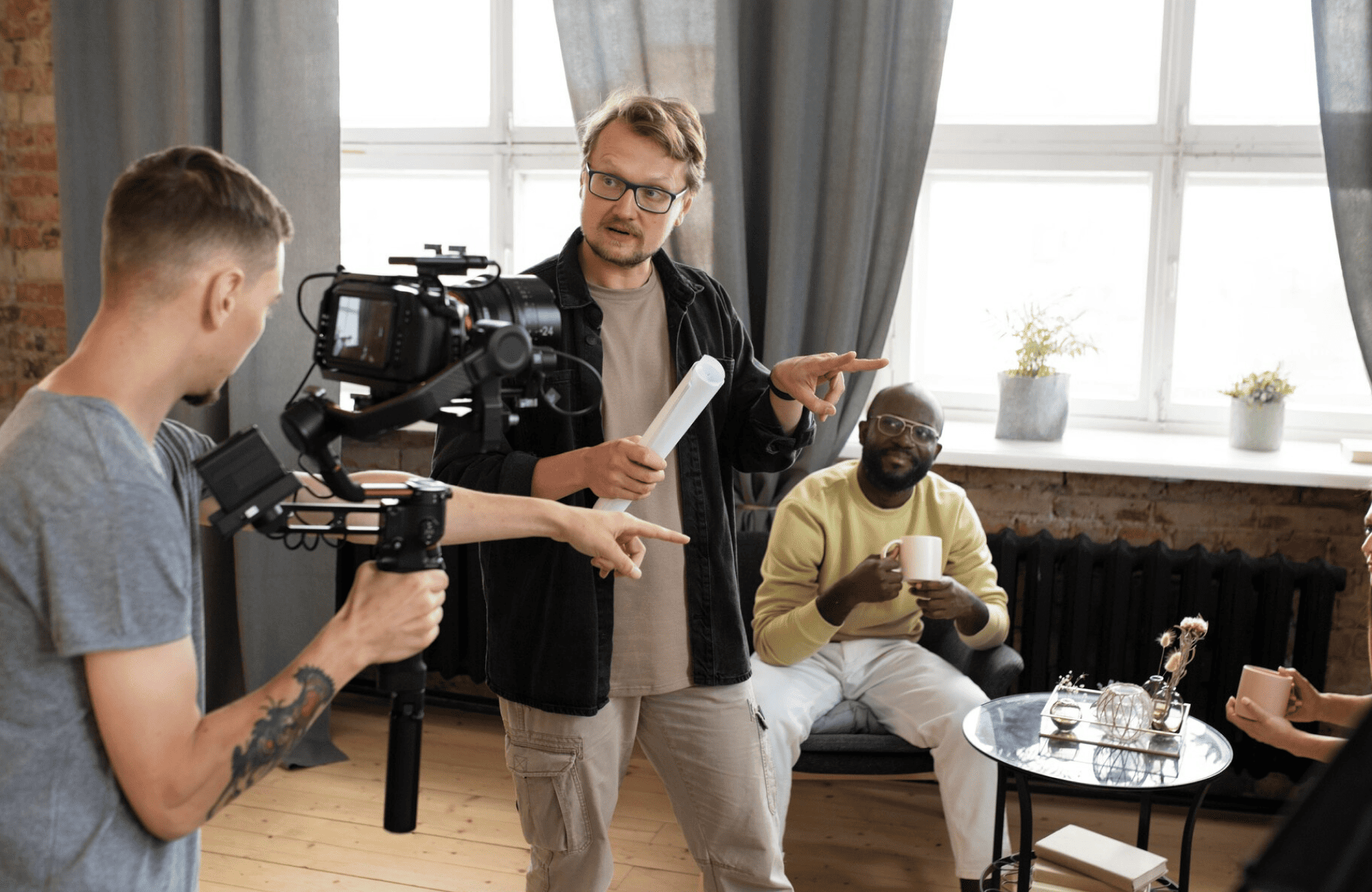 FX Productions Canada A man holding papers gestures while speaking to a cameraman filming him; another man sits on a couch holding a mug, with a table and decor in the foreground—capturing the creative energy of an FX Productions Canada set. Toronto Video Production Company