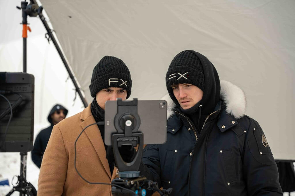 FX Productions Canada Two people wearing black FX beanies and winter coats look at a tablet mounted on a tripod, with film equipment in the background, showcasing the behind-the-scenes work of Toronto Video Production experts. Toronto Video Production Company