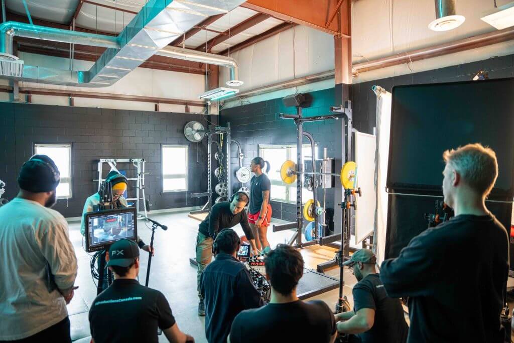 FX Productions Canada A Toronto Video Production crew sets up equipment and records a scene with a person lifting weights in a well-lit gym filled with various fitness equipment. Toronto Video Production Company