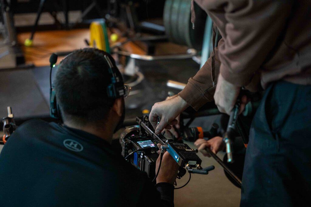 FX Productions Canada Two people wearing headsets adjust settings on a professional video camera in a gym, showcasing Toronto Video Production expertise by FX Productions Canada, with gym equipment visible in the background. Toronto Video Production Company