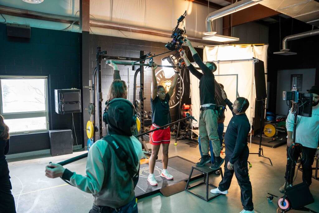 FX Productions Canada A film crew from Toronto Video Production sets up cameras and boom microphones around a person in sports attire standing in a gym with weightlifting equipment. Toronto Video Production Company