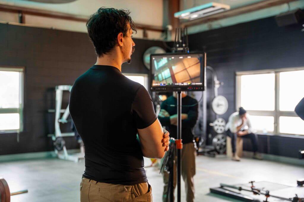 FX Productions Canada A man in a black shirt stands with arms crossed, looking at a monitor in a gym setting with equipment and large windows, capturing the behind-the-scenes vibe of a Toronto Video Production shoot. Toronto Video Production Company