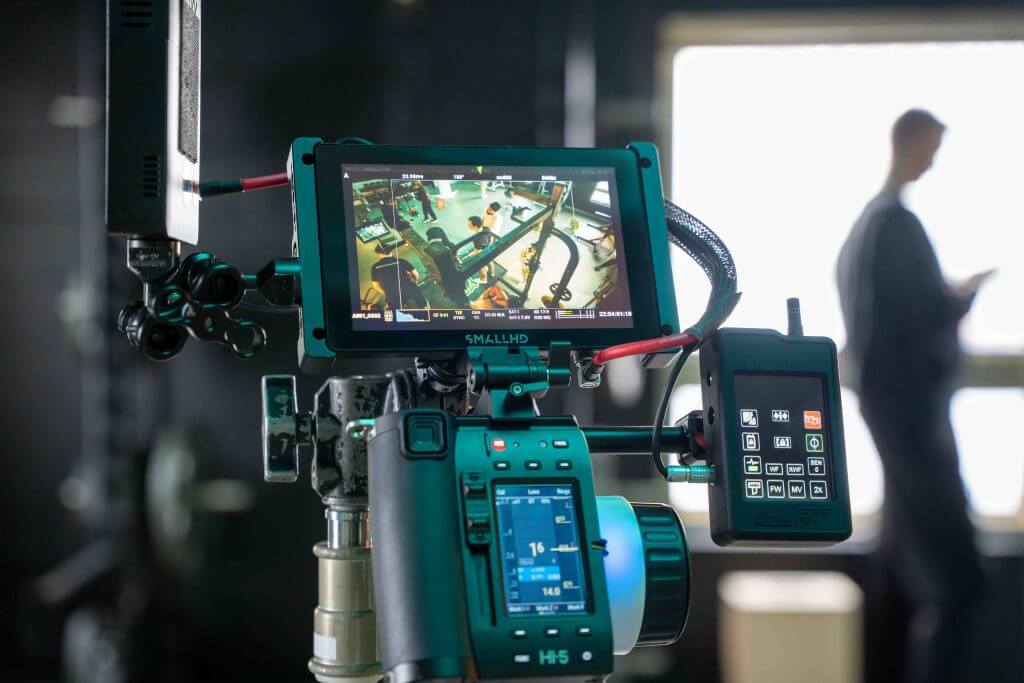FX Productions Canada A professional video camera from a Toronto Video Production company displays a live feed of a film set with crew members, while a person stands blurred in the background near a window, using a phone. Toronto Video Production Company