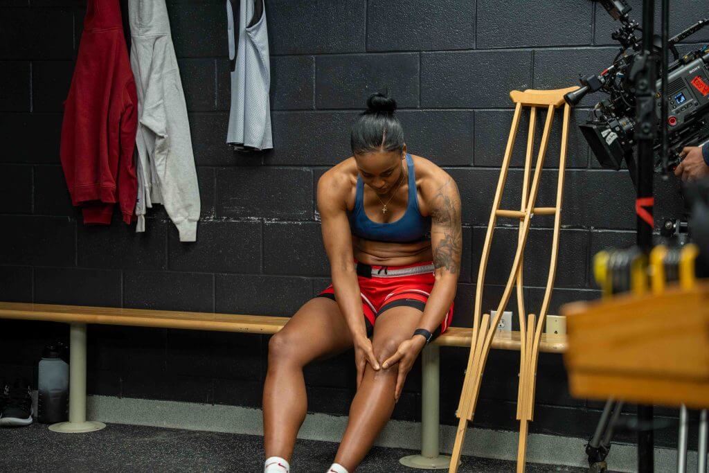 FX Productions Canada A woman in athletic wear sits on a bench in a locker room, touching her leg. Crutches and hanging clothes are nearby, with a Toronto Video Production crew filming her. Toronto Video Production Company