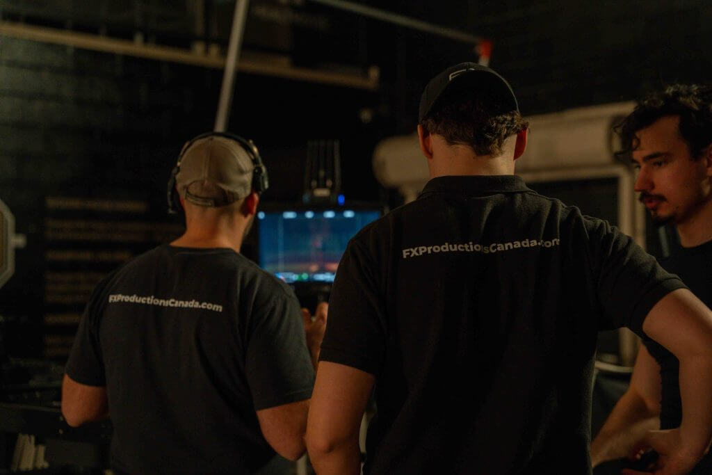 FX Productions Canada Three crew members stand in a dimly lit room, two wearing shirts with "FXProductionsCanada.com" on the back, as they review footage on a monitor for Toronto Video Production. Toronto Video Production Company