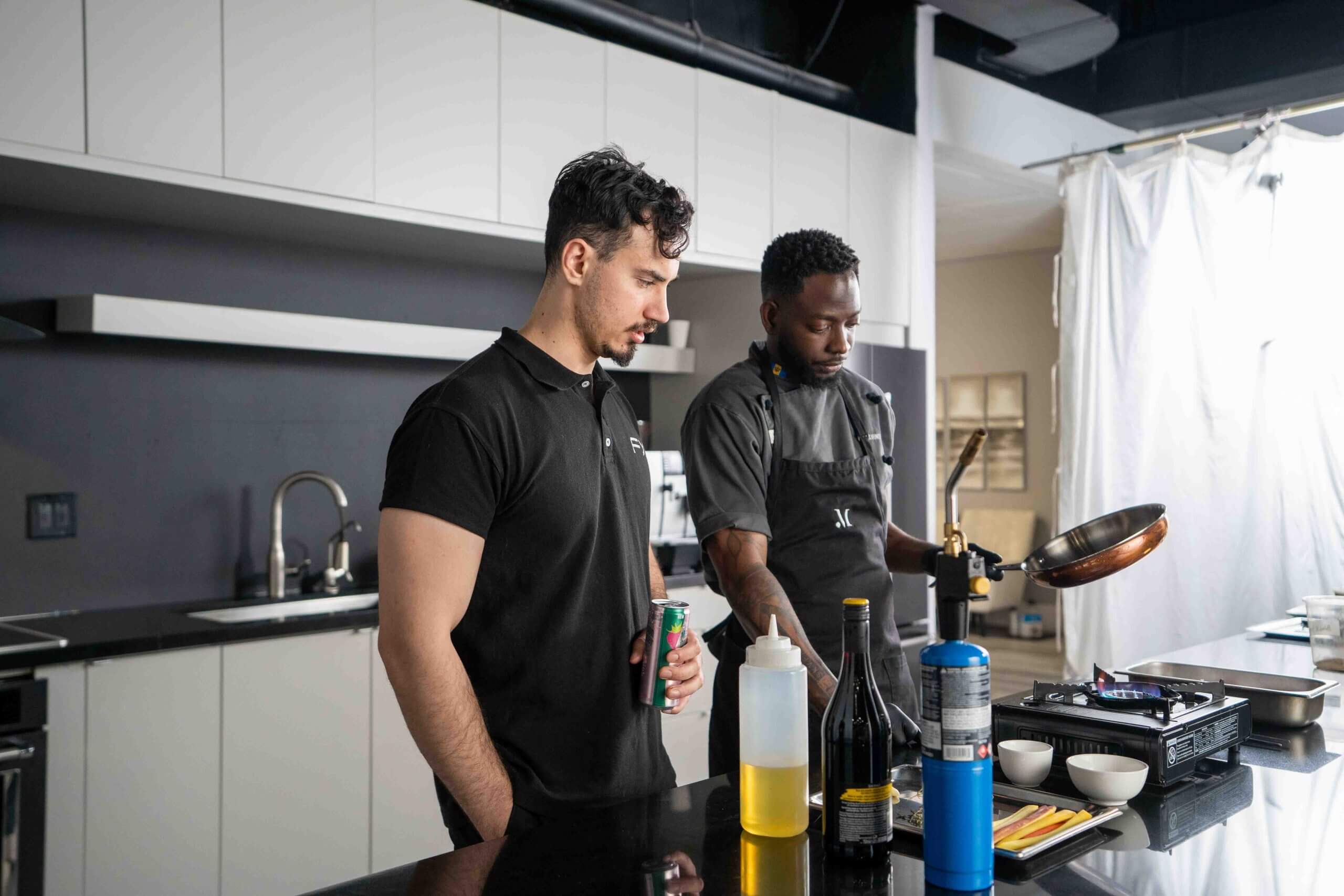 FX Productions Canada Two men stand in a modern kitchen, one holding a drink and the other cooking at a counter with oil, bottles, and kitchen tools—an ideal setting for an FX Productions Canada video production. Toronto Video Production Company