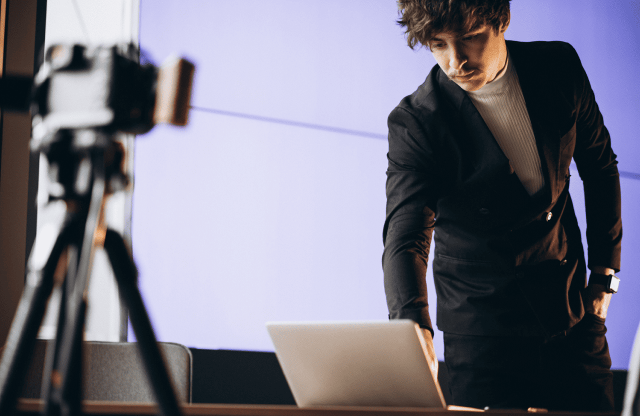 FX Productions Canada A person in a black suit adjusts a laptop on a desk in front of a camera tripod, with a purple background behind them, creating content for FX Productions Canada. Toronto Video Production Company