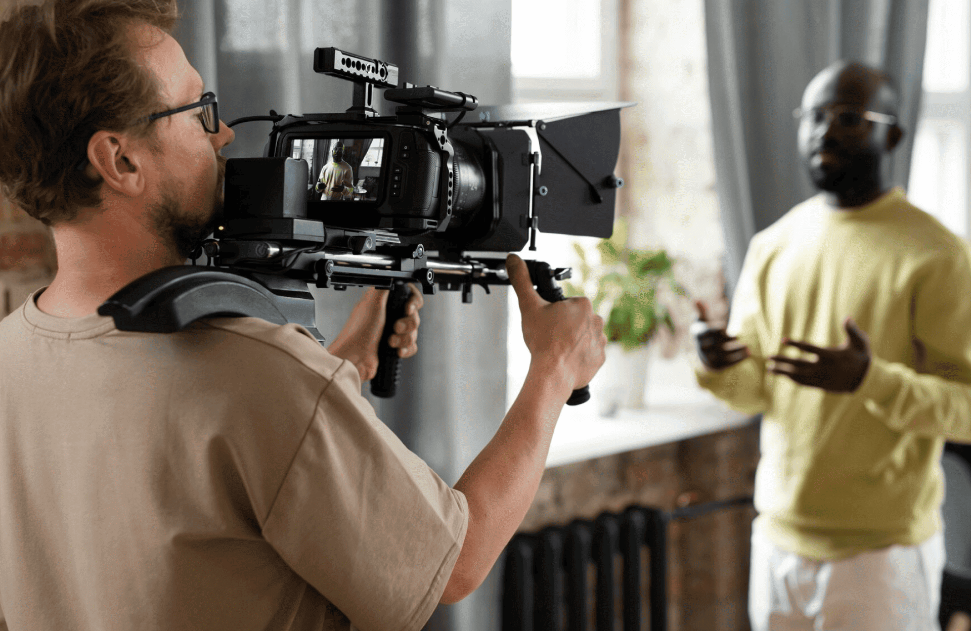 FX Productions Canada A cameraman films a man speaking indoors. The speaker stands in front of a window and gestures with his hands while the cameraman operates professional video equipment. Toronto Video Production Company