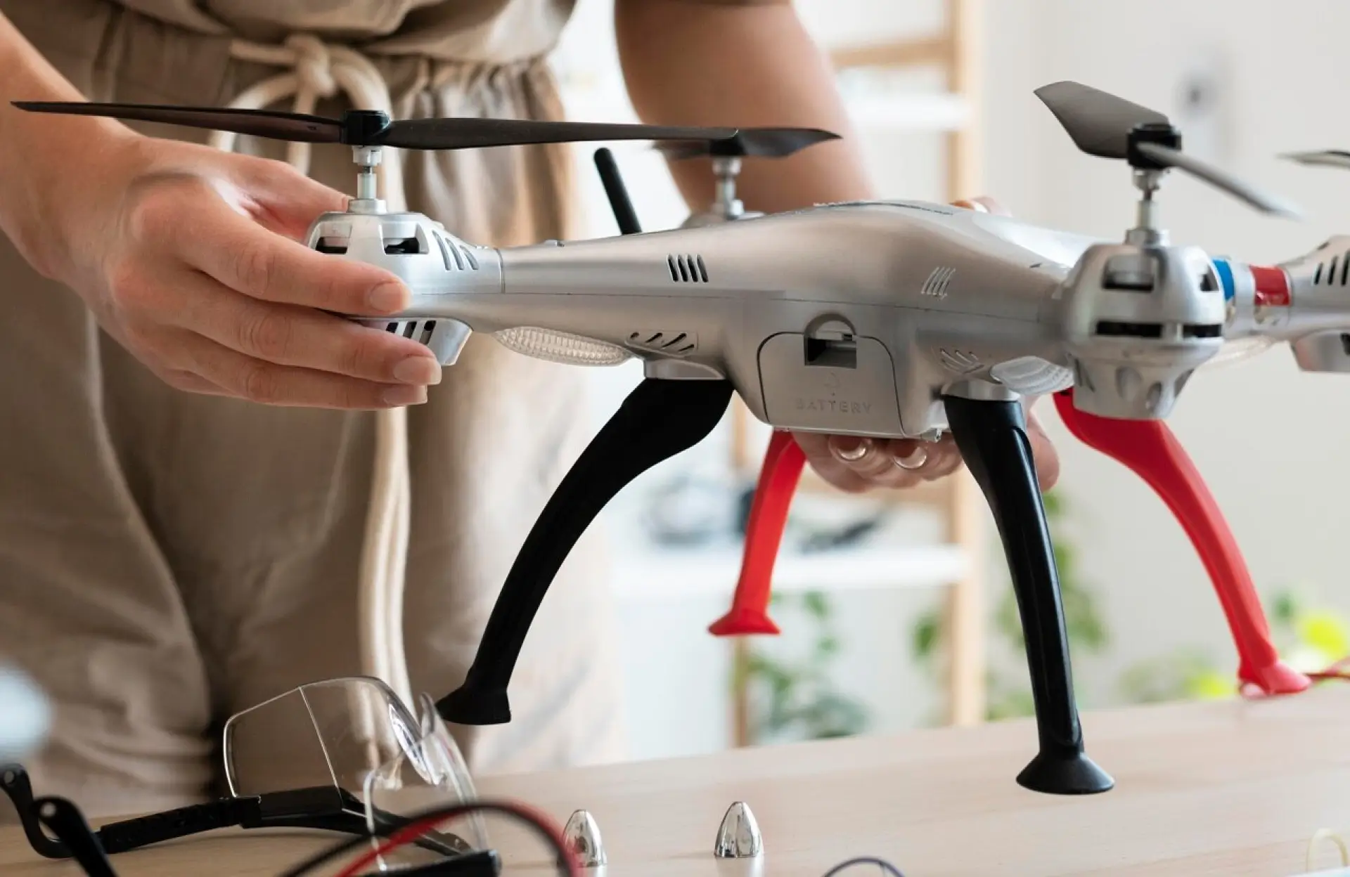 FX Productions Canada A person holds a silver quadcopter drone with black and red landing gear, standing indoors on a wooden table with drone parts nearby. Toronto Video Production Company