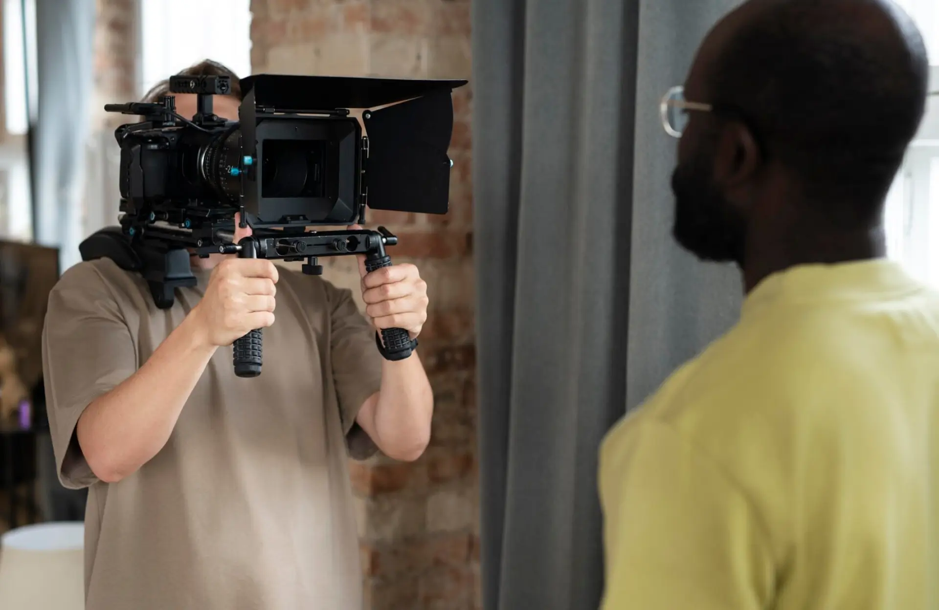 FX Productions Canada A person operates a professional video camera while filming another person indoors, with a curtain and brick wall in the background. Toronto Video Production Company