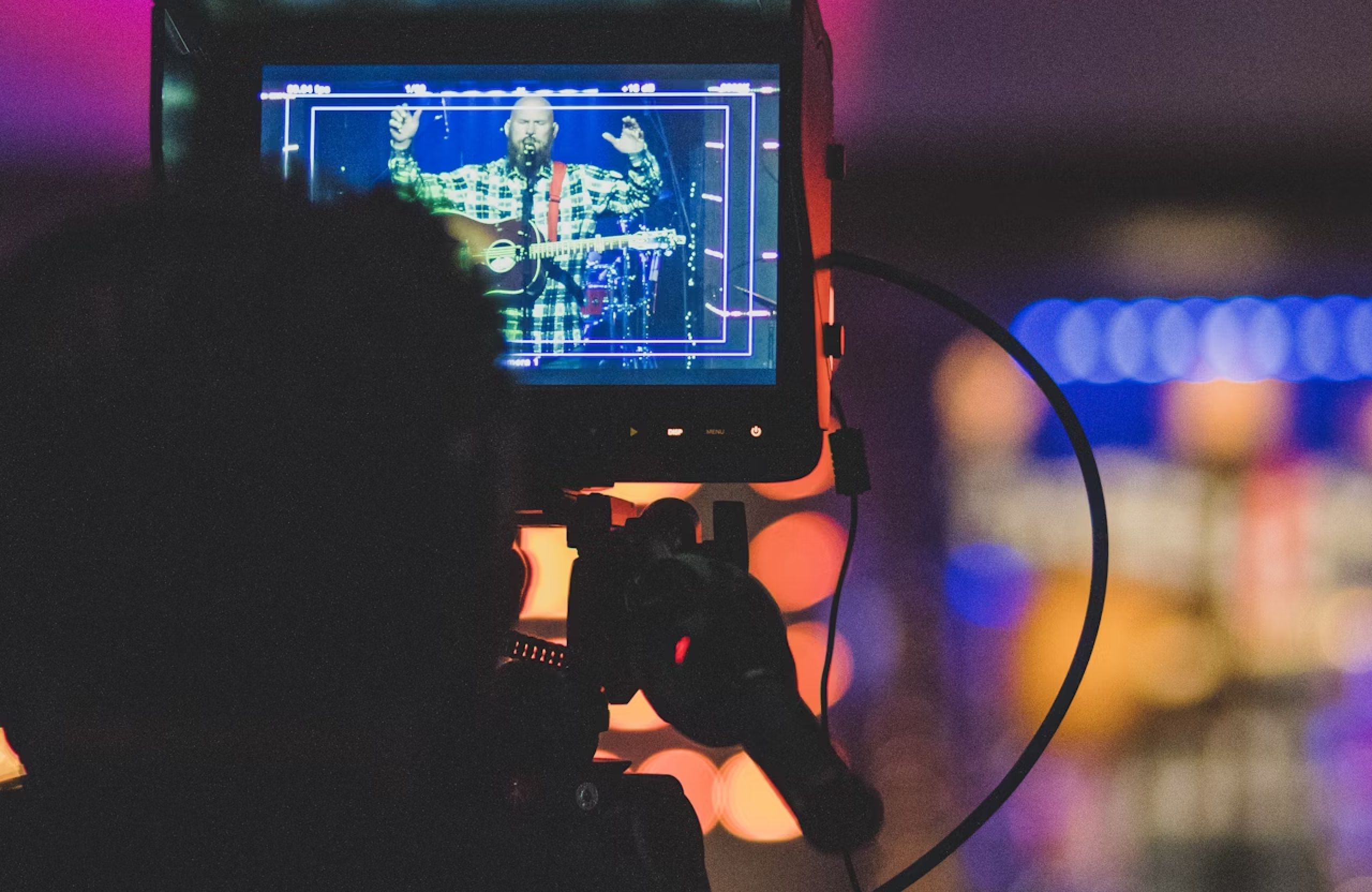 FX Productions Canada A camera films a musician playing guitar and singing on stage, as seen through the camera's monitor with colorful blurred lights in the background—a perfect scene for Toronto music video production. Toronto Video Production Company