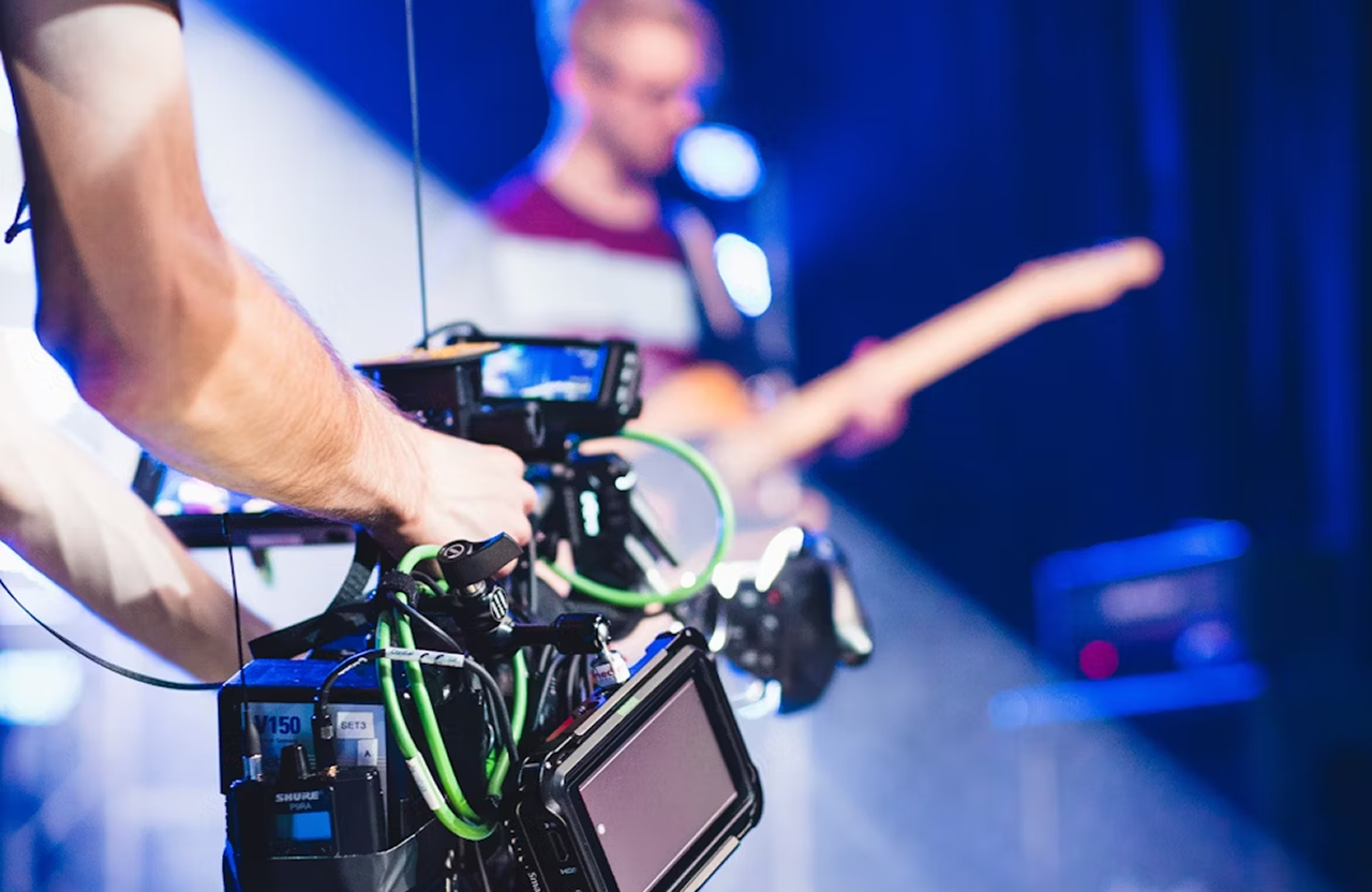 FX Productions Canada A person operates a professional video camera while a musician plays an electric guitar on stage in the background, capturing the energy of cinematic video production. Toronto Video Production Company
