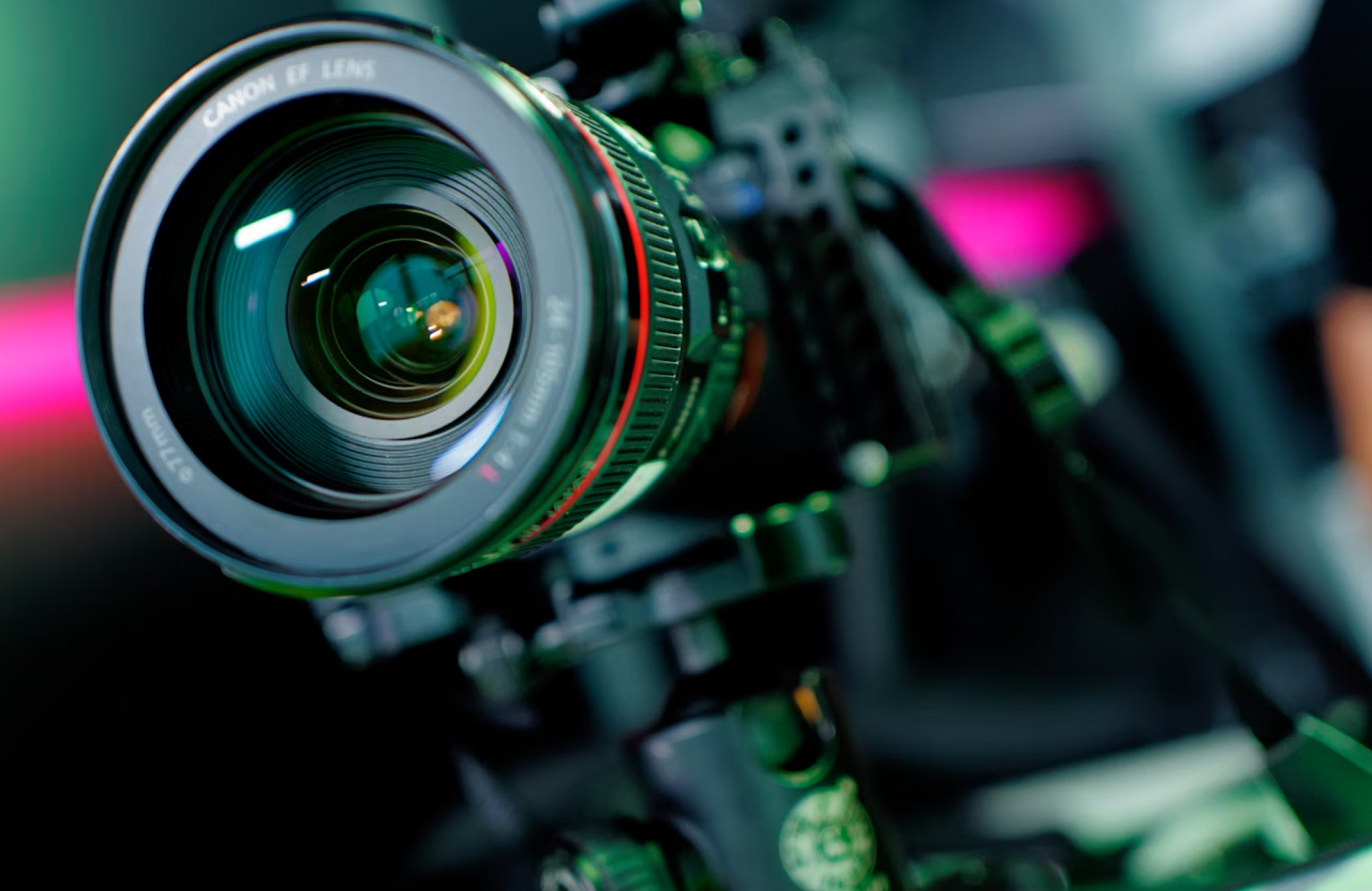 FX Productions Canada Close-up of a professional Canon camera with an EF lens mounted on a tripod, shown in sharp focus against a blurred background with pink and green light accents. Toronto Video Production Company