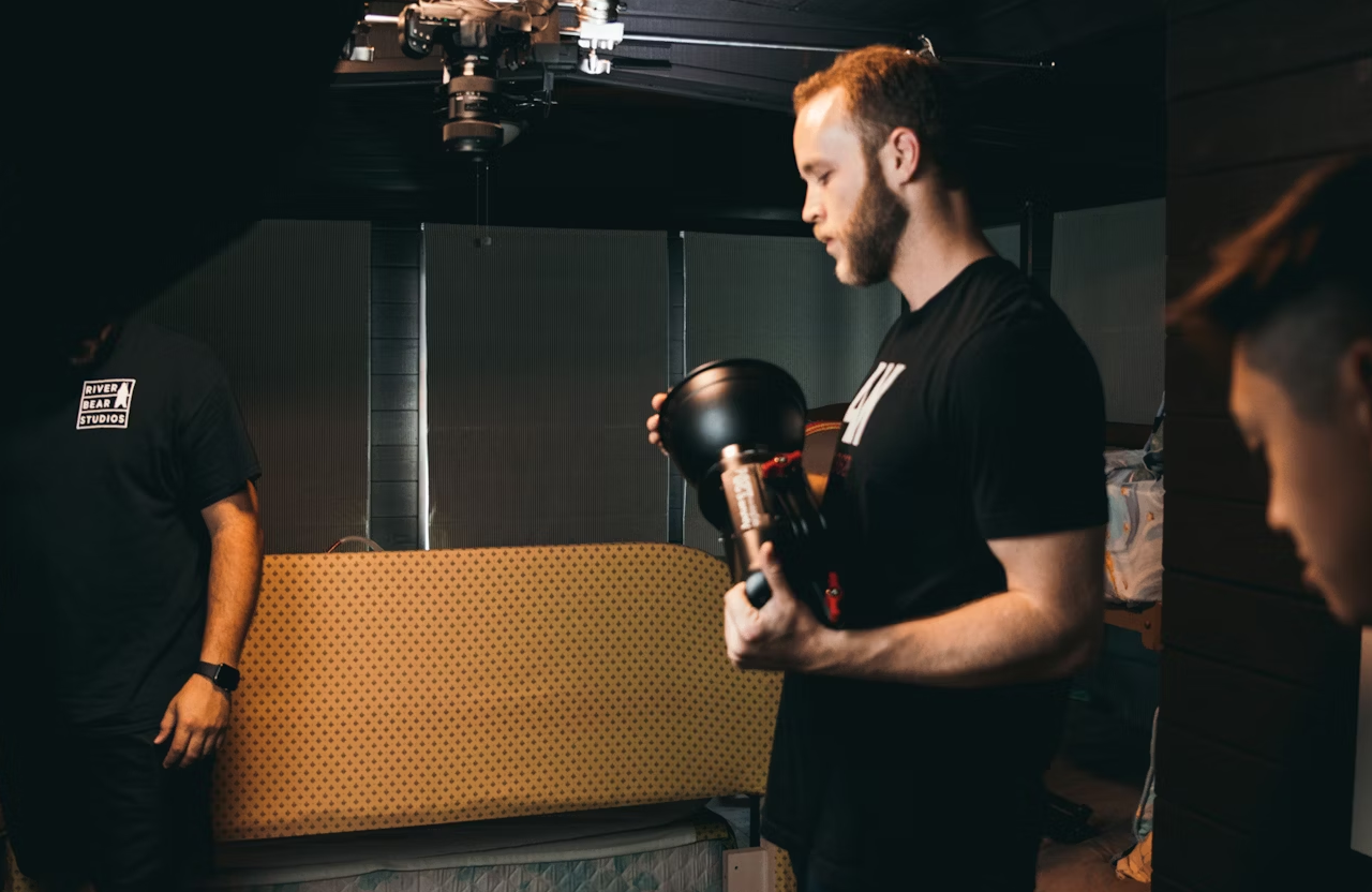FX Productions Canada A man in a black t-shirt holds camera equipment in a dimly lit room with a patterned couch and a ceiling-mounted light above. Toronto Video Production Company