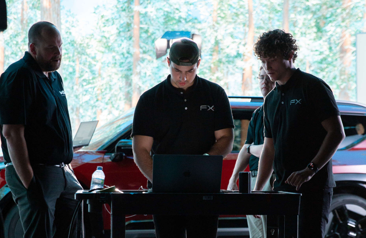 FX Productions Canada Three men in black FX shirts stand around a table, working on a laptop indoors, with a red car and trees visible in the background. Toronto Video Production Company