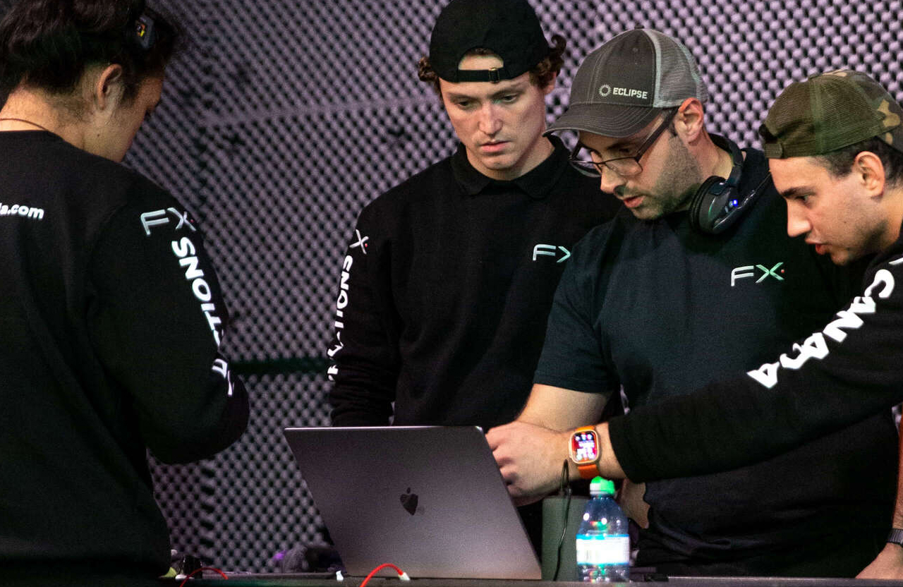 FX Productions Canada Four people stand around a table, focused on a laptop. One person points at the screen while others observe. A water bottle and headphones are also on the table. Toronto Video Production Company