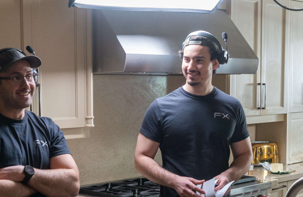 FX Productions Canada Two men wearing headsets and FX-branded shirts stand and talk in a kitchen with light-colored cabinets and a stove. Toronto Video Production Company