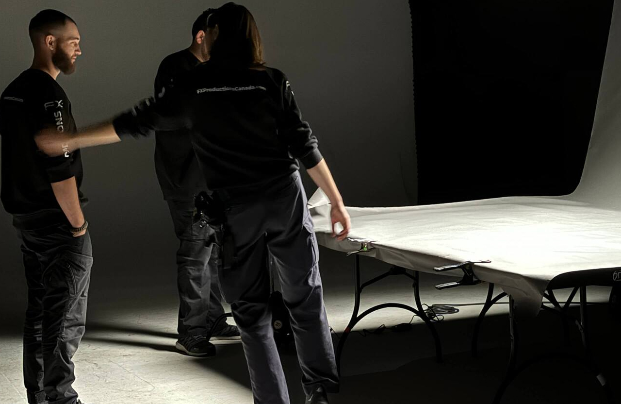 FX Productions Canada Three people wearing dark clothing stand around a table covered with a white sheet in a studio with a dark background and professional lighting. Toronto Video Production Company