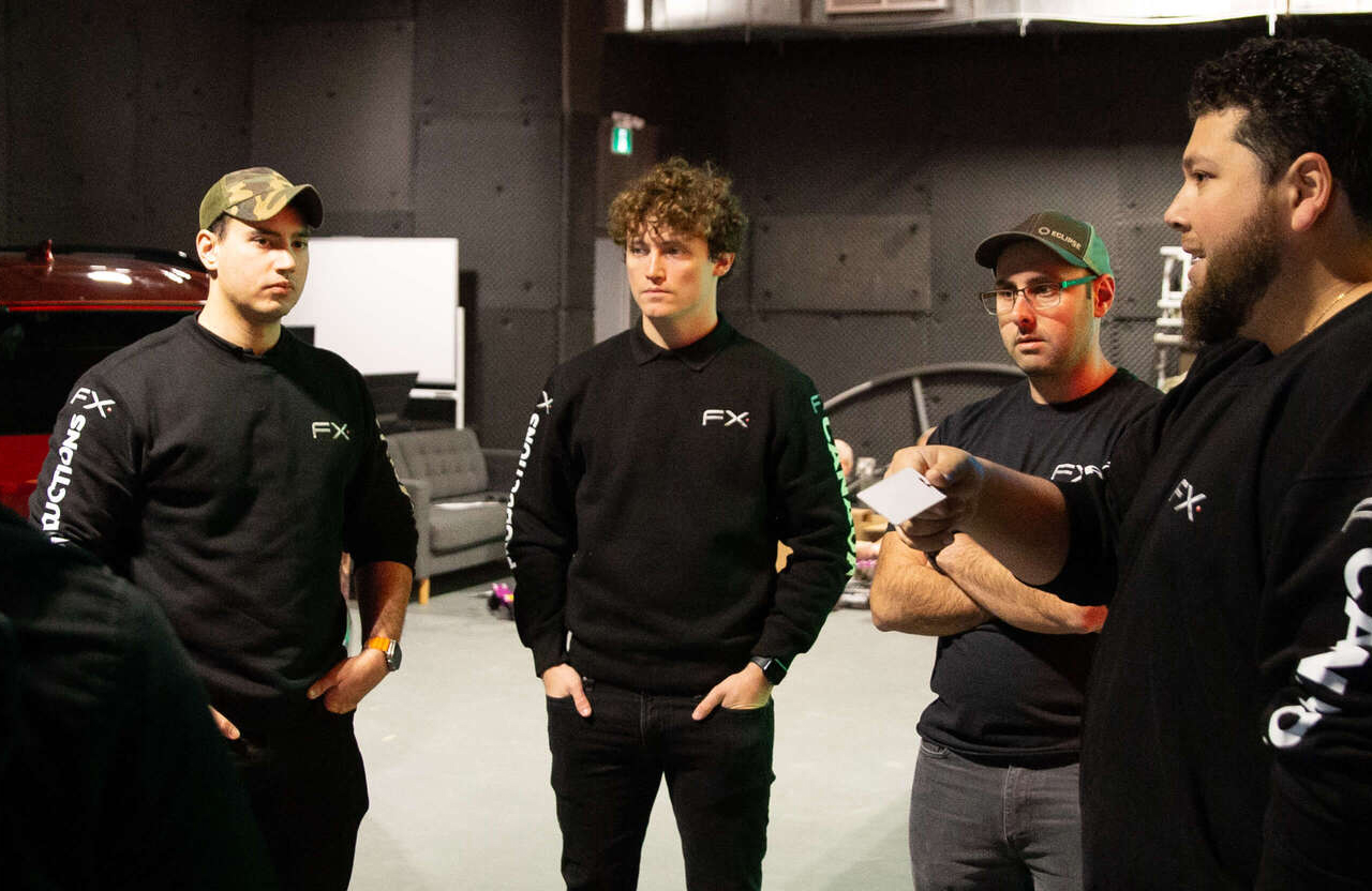 FX Productions Canada Four men wearing black FX sweaters stand in a group, listening to another person who is holding a piece of paper in a dimly lit room with equipment in the background. Toronto Video Production Company