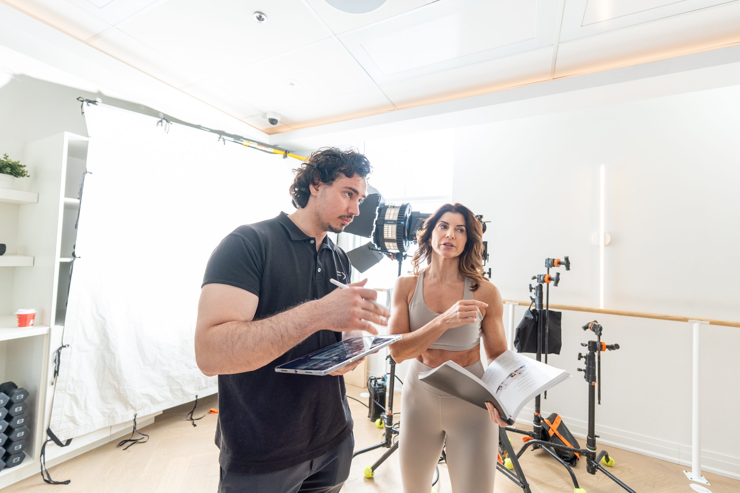 FX Productions Canada A man holding a laptop and a woman holding papers have a discussion in a brightly lit studio with lighting equipment in the background. Toronto Video Production Company