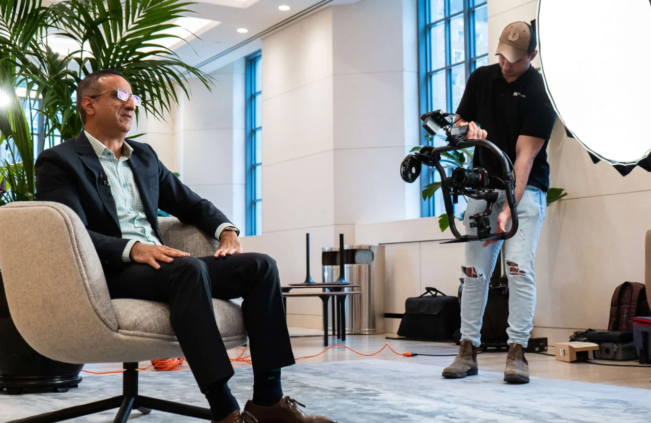 FX Productions Canada A man in a suit sits in a chair while a videographer adjusts camera equipment, capturing an executive corporate video production in a modern, well-lit room with large windows. Toronto Video Production Company