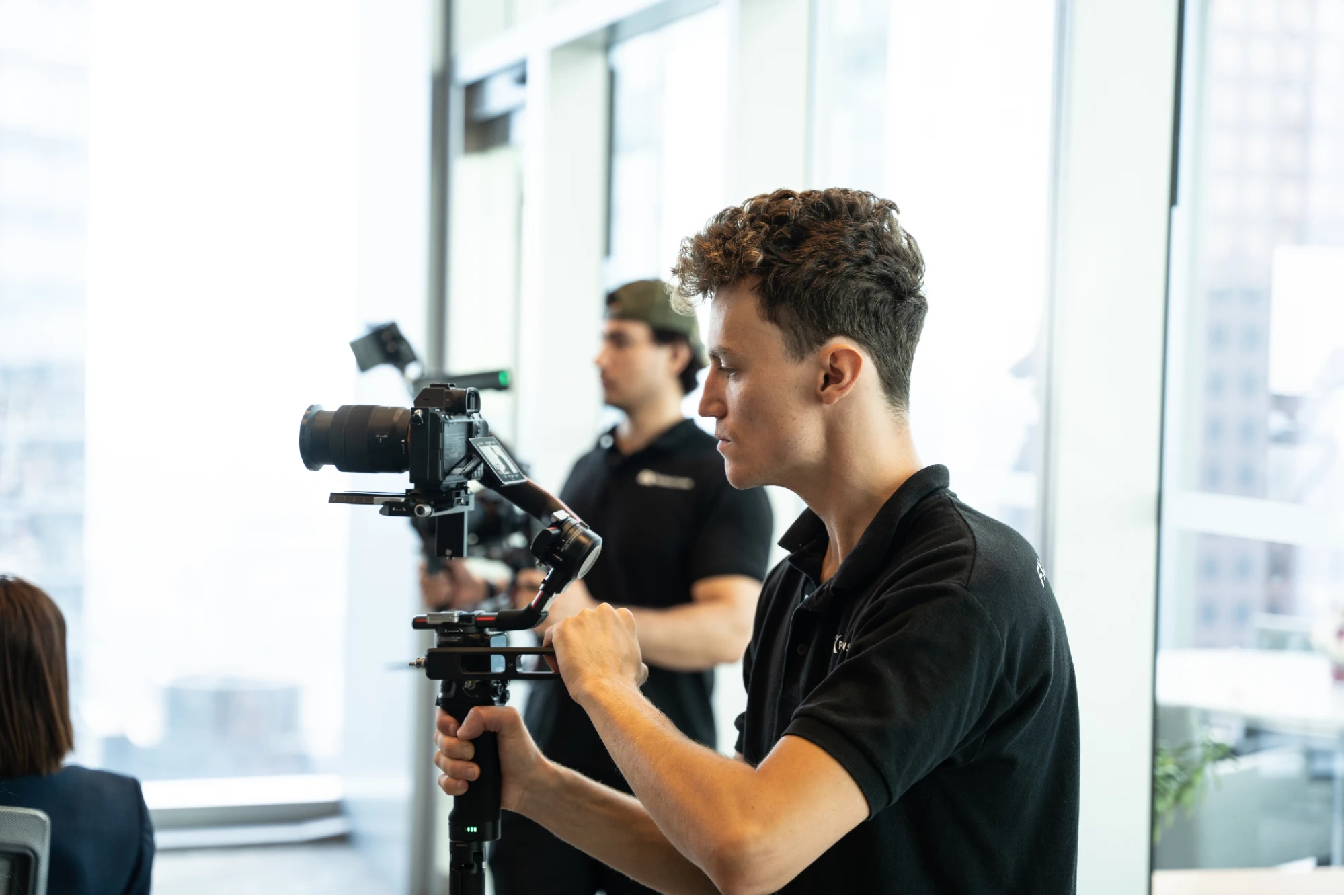 FX Productions Canada A man operates a camera mounted on a stabilizer in a modern, well-lit indoor setting, showcasing executive corporate video production with another person and filming equipment visible in the background. Toronto Video Production Company