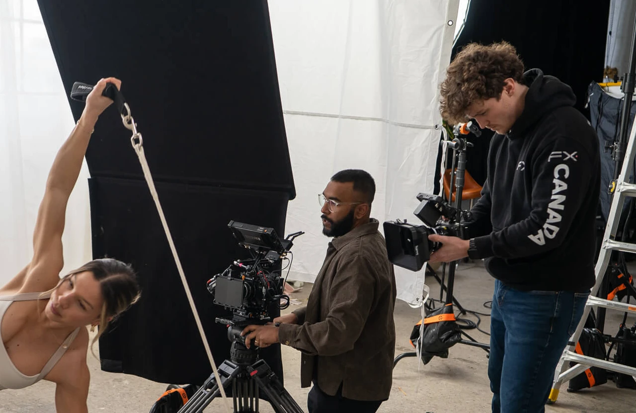 FX Productions Canada A woman exercises with resistance bands while two men film her using professional camera equipment in a studio setting. Toronto Video Production Company