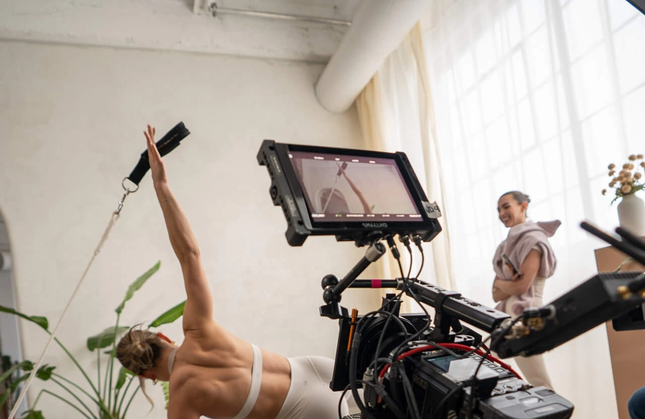 FX Productions Canada A camera films a woman stretching with a resistance band, while another woman stands in the background near a large window with sheer curtains. Toronto Video Production Company