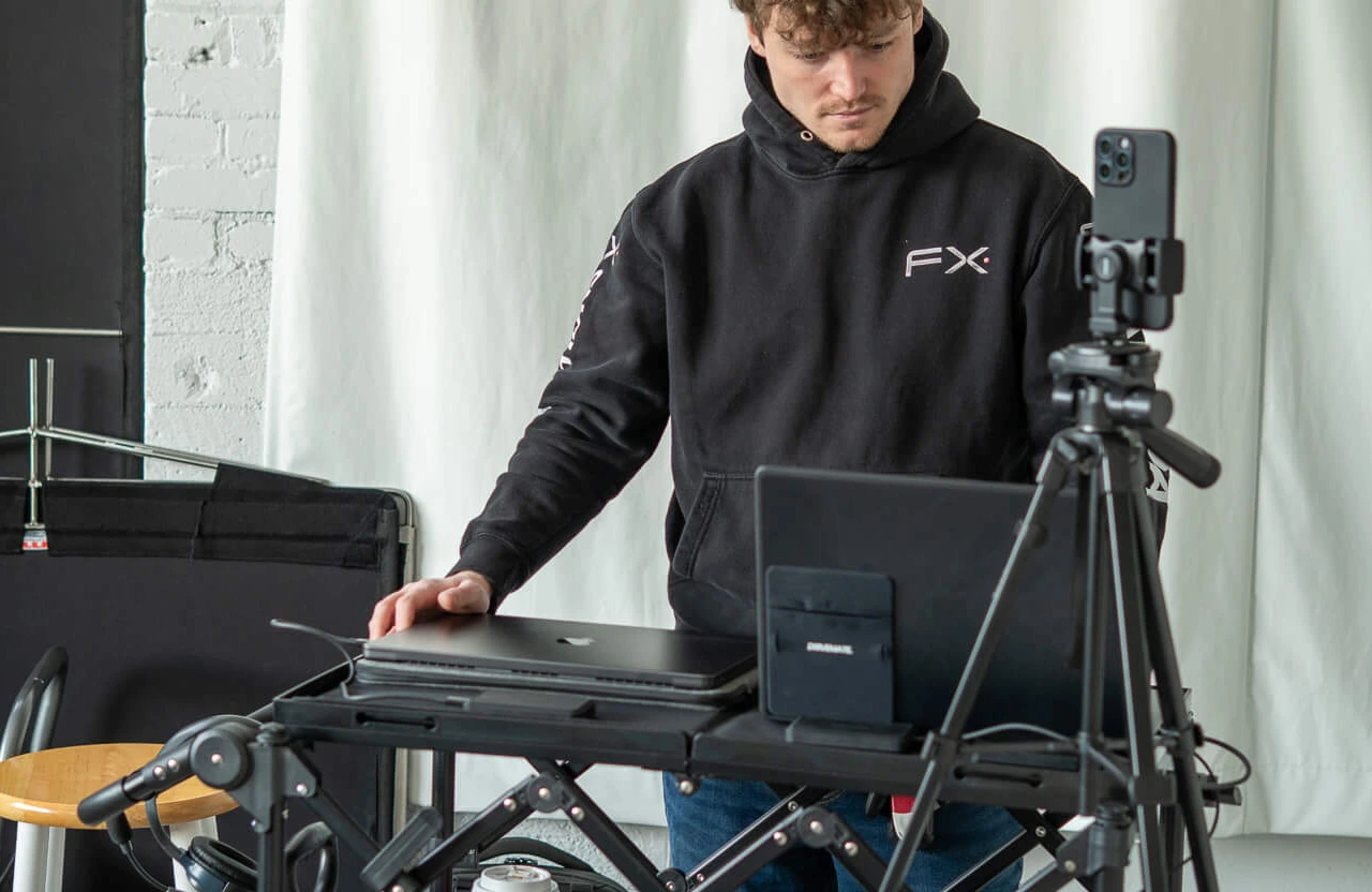 FX Productions Canada A person in a black hoodie stands at a desk with a laptop, smartphone on a tripod, and other electronics, appearing to set up or operate recording equipment for a creative production company Toronto. Toronto Video Production Company
