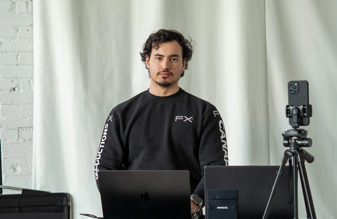 FX Productions Canada Man with dark hair and a black sweatshirt sits at a desk with two laptops and a smartphone on a tripod, against a light curtain background, working on automotive video production Toronto projects. Toronto Video Production Company