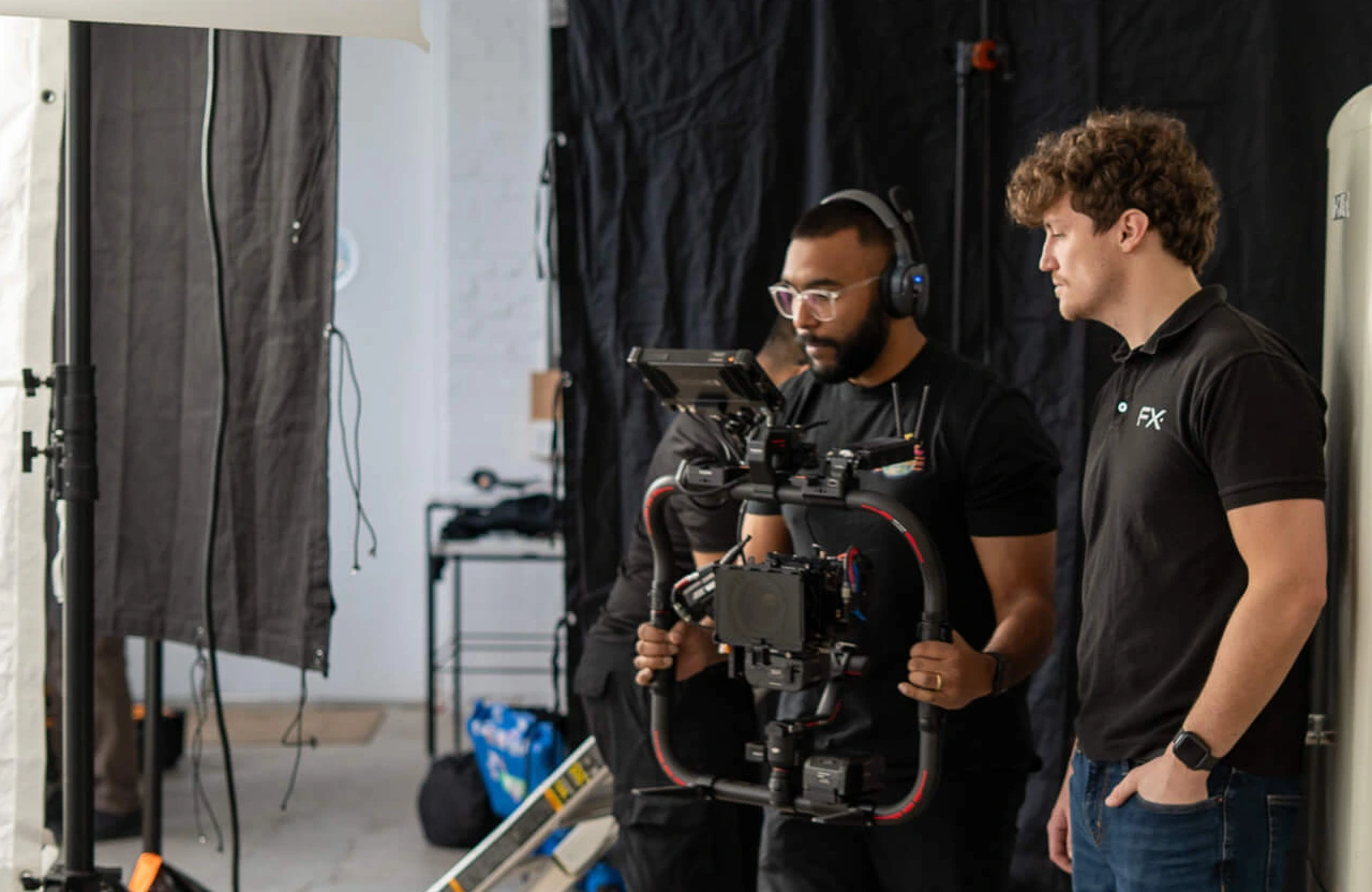 FX Productions Canada Two men stand in a studio; one operates a professional camera rig while the other observes. Black curtains and equipment are visible in the background. Toronto Video Production Company