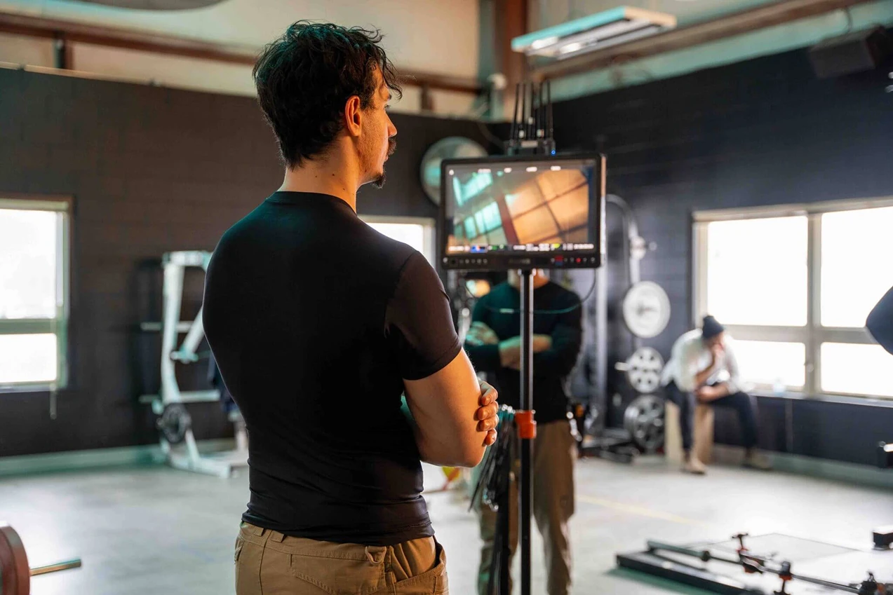 FX Productions Canada A man stands with arms crossed, looking at a monitor in a gym setting, while another person sits by the windows in the background. Toronto Video Production Company