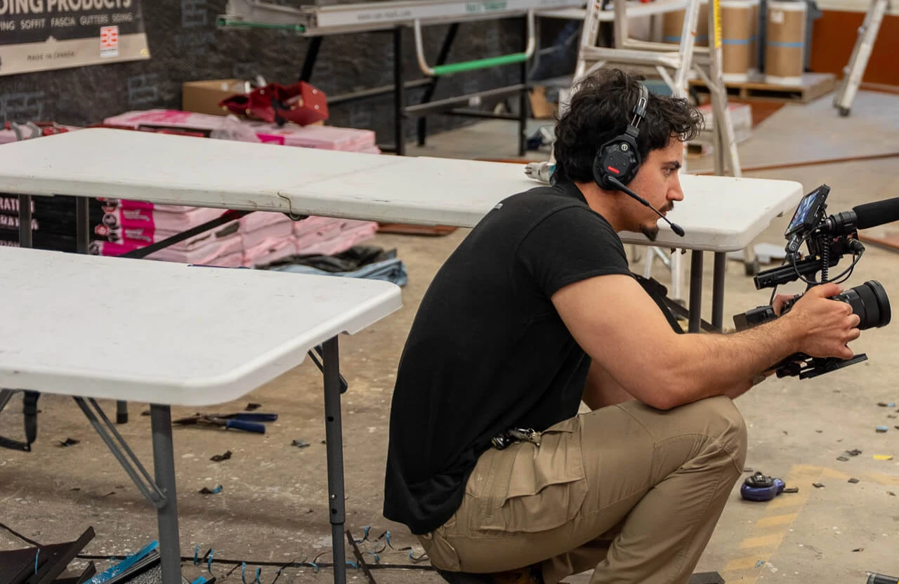 FX Productions Canada A man wearing a headset crouches next to folding tables, holding a professional video camera in a workshop or warehouse setting. Toronto Video Production Company